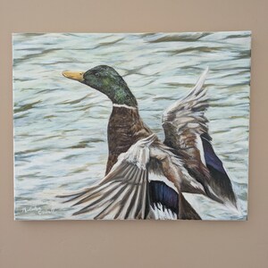 May include: Oil painting of a mallard duck with outstretched wings, set against a backdrop of water. The duck features a green head, brown body, and gray and blue wings. The artwork is signed in the lower left corner.