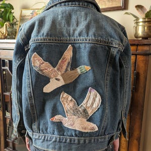 May include: A denim jacket with two embroidered duck patches on the back. The ducks are depicted in flight with detailed wing patterns and colorful accents. The jacket is a medium wash blue.