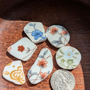 May include: Seven pieces of vintage porcelain with floral designs in shades of blue, orange, and gold. The pieces are irregularly shaped and arranged on a wooden surface. A quarter is included for size reference.