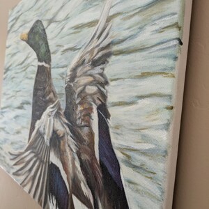 May include: A canvas painting depicts a duck with outstretched wings, set against a backdrop of water. The duck features a green head, brown body, and gray and blue wings. The artwork is rendered with visible brushstrokes, creating a textured effect.