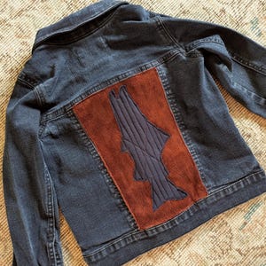 May include: Dark blue denim jacket with a brown suede patch on the back. The patch features a dark blue, quilted design. The jacket has a classic collar, buttoned cuffs, and a button closure.