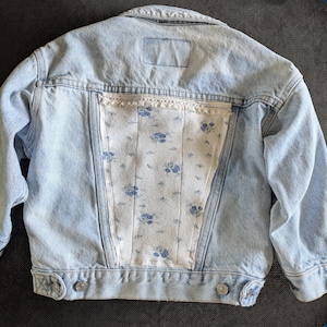 Puede incluir: Chaqueta vaquera azul claro con un panel trasero estampado floral. El panel trasero es blanco con diseños florales azules y está adornado con encaje. La chaqueta tiene cierres de botón y dos bolsillos delanteros.