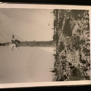 May include: Black and white photograph of a tall column with figures at the top, possibly a monument or statue. The background shows a cityscape with buildings and what appears to be a crowd of people. The image is in a protective plastic sleeve.