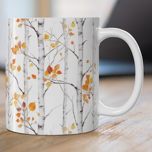 May include: White ceramic mug featuring a watercolor design of birch trees with orange and yellow leaves. The mug has a curved handle and is set on a wooden surface, with a laptop in the background.