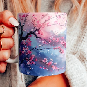 May include: White ceramic mug featuring a watercolor design of a cherry blossom tree with pink flowers against a blue and pink sky. The mug has a white handle and is held by a person.