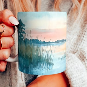 May include: A white ceramic mug with a watercolor design of a lake scene with tall grass and a sunset sky.