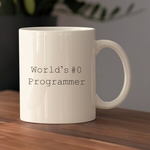 Könnte beinhalten: Eine weiße Keramiktasse mit einem schwarzen Textdesign, das "World's #0 Programmer" lautet.