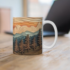 May include: White ceramic mug featuring a layered landscape design with a mountain range, trees, and a sunset in shades of orange, brown, and blue. The mug has a curved handle and is set on a wooden surface.