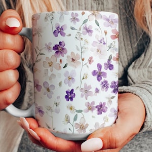 May include: A white ceramic mug with a floral pattern featuring purple and pink flowers. The mug is held by a person's hands.