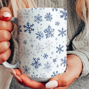 Puede incluir: Taza de cerámica blanca con un diseño de copos de nieve invernales en tonos azules y grises. La taza tiene un asa blanca y se sostiene con ambas manos. El diseño presenta copos de nieve de varios tamaños.