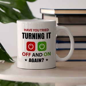 Könnte beinhalten: Weiße Keramiktasse mit dem Text "HAVE YOU TRIED TURNING IT OFF AND ON AGAIN?" in Schwarz, Rot und Grün. Die Tasse zeigt Symbole für Ein-/Ausschalter und Sterne. Die Tasse steht auf einer weißen Oberfläche.