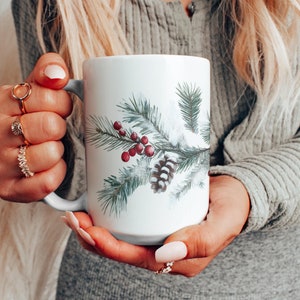 May include: White ceramic mug featuring a winter design with pine needles, red berries, and a pine cone. The mug is held by a person wearing a gray long-sleeved shirt. The mug is a standard size for coffee or tea.