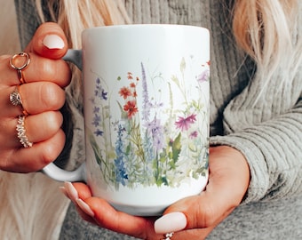 Può includere: Una tazza in ceramica bianca con un design floreale che presenta una varietà di fiori colorati e foglie verdi. La tazza ha un manico ed è tenuta dalle mani di una persona.