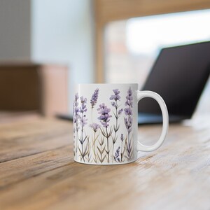 May include: White ceramic mug featuring a watercolor design of lavender flowers in shades of purple and green. The mug has a curved handle and is set on a wooden table. A laptop is visible in the background.