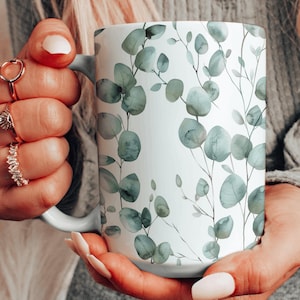 Puede incluir: Taza de cerámica blanca con un diseño de acuarela de hojas de eucalipto verdes.