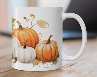 Può includere: Tazza in ceramica bianca con un'illustrazione colorata di zucche e foglie autunnali. Le zucche sono arancioni e bianche e sono disposte in un gruppo.