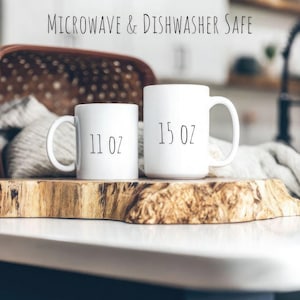 May include: Two white ceramic mugs on a wooden surface. The smaller mug is 11 oz and the larger mug is 15 oz. Both mugs are microwave and dishwasher safe.
