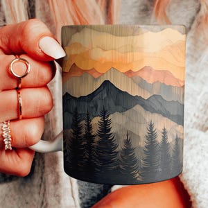 Op de afbeelding: Een witte keramische mok met een houtnerfdesign met een bergketen met een zonsondergangshemel. De bergen zijn donkerblauw en de hemel is een gradiënt van oranje, geel en roze.