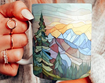 Könnte beinhalten: Eine weiße Keramiktasse mit einem bunten Glasmalereidesign aus einer Bergkette und Kiefern. Das Design enthält Farbtöne von Blau, Grün, Gelb und Lila.