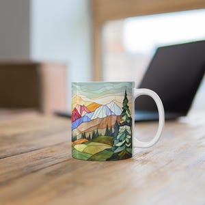 May include: White ceramic mug featuring a colourful stained glass design of a mountain landscape. The design includes a blue sky, green trees, and brown mountains with a touch of red.