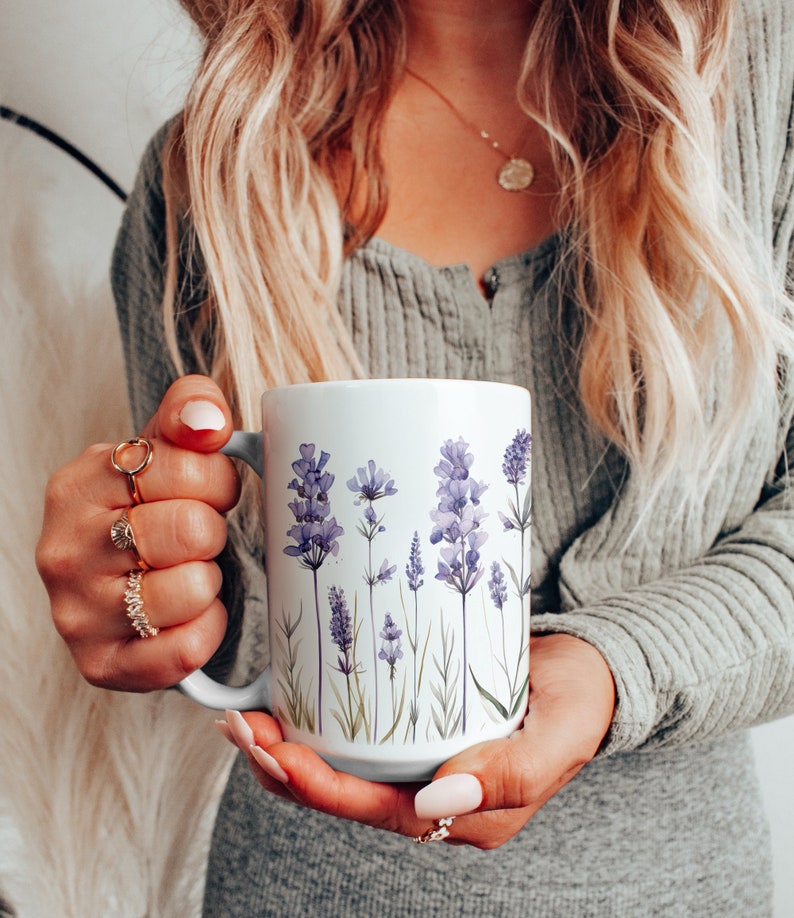 May include: White ceramic mug featuring a lavender floral design. The mug has a light blue handle and is decorated with watercolor illustrations of purple lavender flowers and green stems. A perfect gift for a tea or coffee lover.