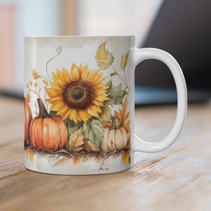 May include: White ceramic mug with a fall-themed design featuring a sunflower and pumpkins. The design is in watercolor style with shades of yellow, orange, brown, and green.