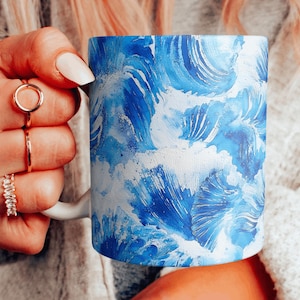 Puede incluir: Taza de cerámica blanca con un diseño de olas en acuarela azul y blanco. La taza está sostenida por una mano que lleva anillos.
