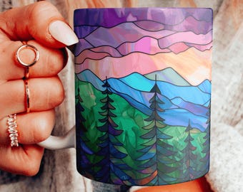 Könnte beinhalten: Eine weiße Keramiktasse mit einem bunten Glasmalereidesign eines Gebirges mit einem Sonnenuntergangshimmel. Das Design zeigt Farbtöne von Lila, Rosa, Blau, Grün und Gelb.