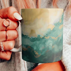 May include: A white ceramic mug with a green and gold abstract design. The design features swirling patterns and hints of yellow.