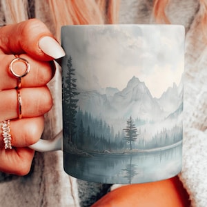 May include: White ceramic mug with a scenic mountain landscape design. The image features a misty mountain range with a lake in the foreground and a forest of pine trees.