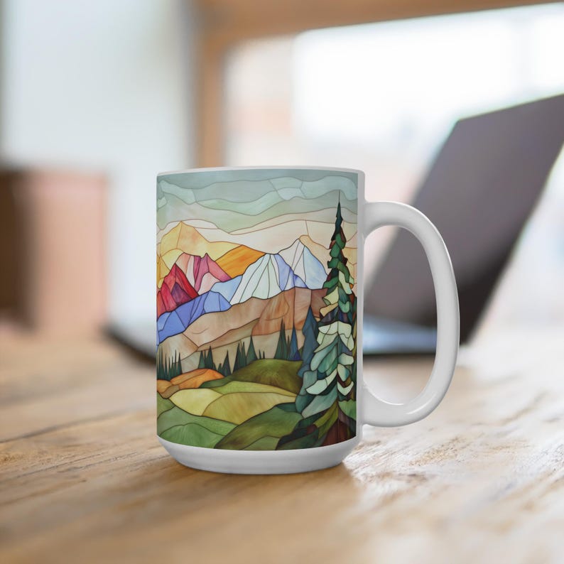 May include: White ceramic mug featuring a colourful stained glass design of a mountain range with a forest in the foreground. The design incorporates shades of blue, green, yellow, orange, and red.