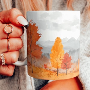 May include: White ceramic mug with a watercolor design of a mountain landscape with trees in shades of orange, yellow, and red. The trees are in the foreground and the mountains are in the background.