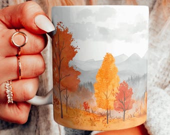 Può includere: Tazza in ceramica bianca con un design ad acquerello di un paesaggio montano con alberi in tonalità di arancione, giallo e rosso. Gli alberi sono in primo piano e le montagne sullo sfondo.