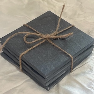May include: A set of four black slate coasters tied together with twine. The coasters are square and have a rustic, natural look.