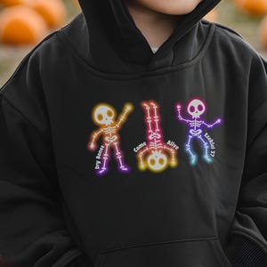 May include: Black hooded sweatshirt featuring three colorful skeleton illustrations with the phrases "Dry Bones", "Come Alive", and "Ezekiel 37". The skeletons are outlined in neon colors, creating a vibrant contrast against the dark fabric.