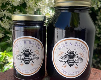 May include: Two glass jars of dark honey with gold lids, labeled "Buzz & Bloom Temecula, CA" with a bee illustration. The jars are set against a backdrop of white flowers and a red brick surface.