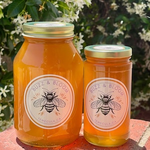 May include: Two glass jars of honey with gold lids and labels that read "Buzz & Bloom Temecula, CA" with a bee illustration. The jars are filled with golden honey and sit on a red brick surface, with a background of green foliage.