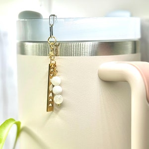 May include: A gold charm with a crescent moon design and three white beads hangs from a silver clip on a light pink and silver travel mug.