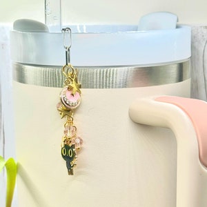 May include: A gold charm with a pink crescent moon and stars, pink beads, and a black cat charm hangs from a silver chain attached to a beige travel mug.