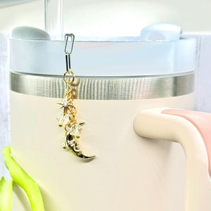 May include: A gold charm with a crescent moon and stars hangs from a silver clip. The charm is attached to a white and silver tumbler.