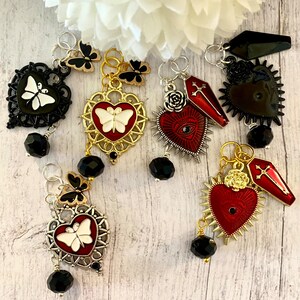 May include: Six charm pendants with a gothic theme. Each charm features a heart with a butterfly, a coffin, and a cross. The charms are made of metal and have black and red enamel. The charms are attached to black bead chains.