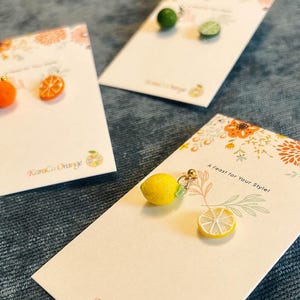 Könnte beinhalten: Drei Karten mit einem floralen Design und dem Text "A Twist for Your Style!" und "Kika Orange" mit kleinen Ohrringen in Form von Orangen- und Zitronenfrüchten auf jeder Karte.