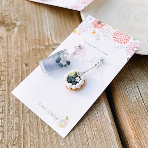 May include: Novelty earrings with miniature food designs. One earring is a purple drink with dark blue spheres, the other is a small tart with white frosting and blueberries. The earrings are on a white card with floral accents and the text "A Feast for Your Style!"