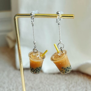 Peut inclure: Une paire de supports de boucles d'oreilles en or avec une base en bois. Chaque boucle d'oreille présente une tasse de boba miniature avec une paille jaune.