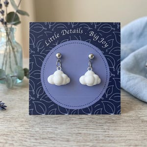 May include: A pair of white cloud-shaped dangle earrings with silver-toned metal posts and jump rings, displayed on a dark blue rectangular card featuring the text Little Details . Big Joy and a white line-art leaf pattern on the background.