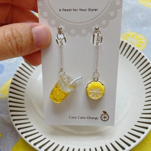 May include: A pair of dangle earrings featuring a lemon slice and a glass of lemonade charm. The earrings are silver and have a chain link design. The card reads "A Feast for Your Style! Cara Cara Orange".