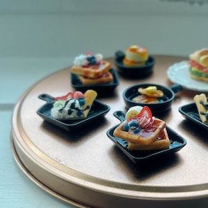 May include: A set of miniature waffle dishes with various toppings, including fruit, whipped cream, and chocolate sauce. The dishes are arranged on a gold-colored tray.