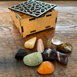 May include: A wooden box with a decorative, openwork lid sits open, next to a collection of polished gemstones. The gemstones are various colors, including black, orange, pink, and clear. The box and gemstones are on a wooden surface.