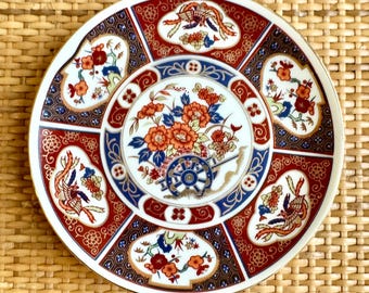 May include: A white ceramic plate with a gold rim and a colorful Japanese-inspired design. The plate features a central image of a cart with flowers and a bird on each side.