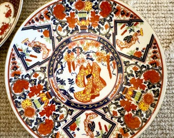 May include: A white ceramic plate with a gold rim and a colorful floral design. The design features red, orange, yellow, blue, and black flowers and leaves. A woman in a red and yellow kimono is depicted in the center of the plate.